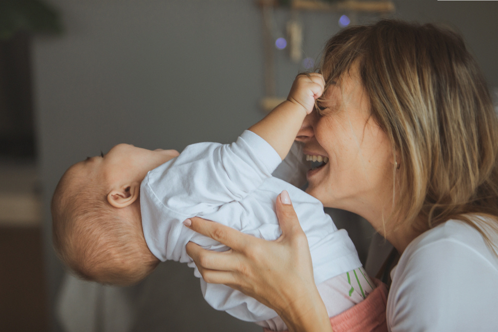 A smiling woman lifts a baby who is laughing happily and touching the woman's forehead with his hand. A warm, intimate moment.